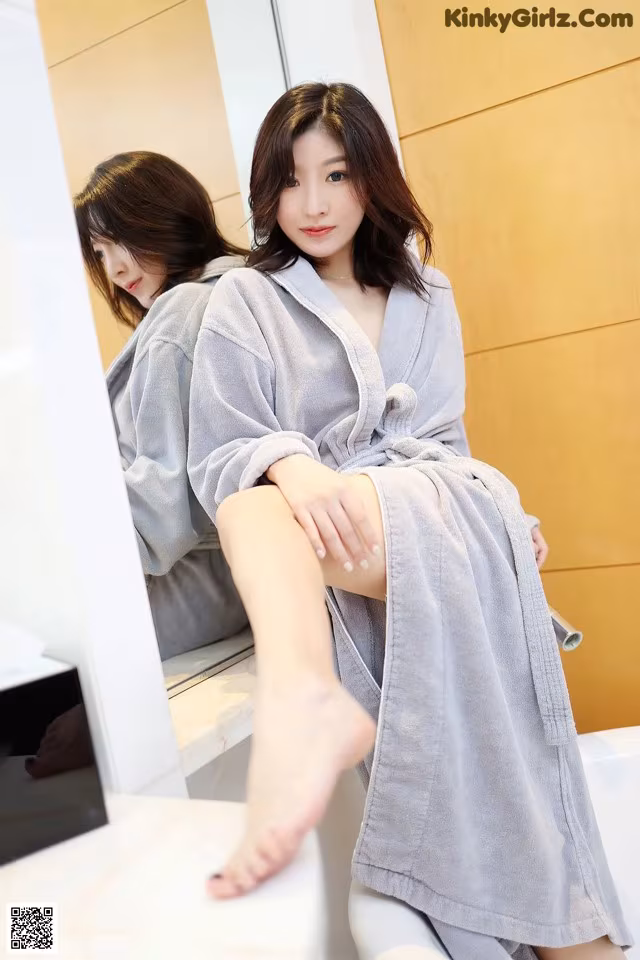 A woman in a bathrobe sitting on a bathtub in front of a mirror.