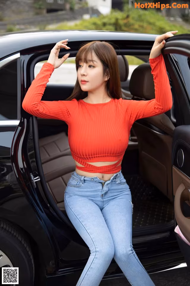 A woman in a red top and jeans leaning out the door of a car.